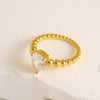 Bague dorée avec perles blanches et petit cœur, posée sur fond blanc