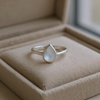Bague délicate en argent sterling avec nacre de perle naturelle en forme de goutte aux reflets irisés subtils, sertissage clos, design minimaliste sur fond blanc
