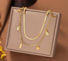 Collier double chaîne dorée avec pendentifs étoiles, éclairs, lunes et œil protecteur, présenté sur un coussin beige.