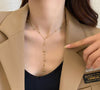Collier en Y doré avec petites perles sphériques, porté sur un haut noir et veste beige.