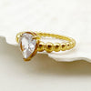 Bague dorée avec perles blanches et petit cœur, posée sur fond blanc