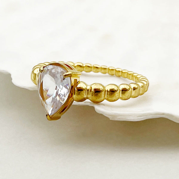 Bague dorée avec perles blanches et petit cœur, posée sur fond blanc
